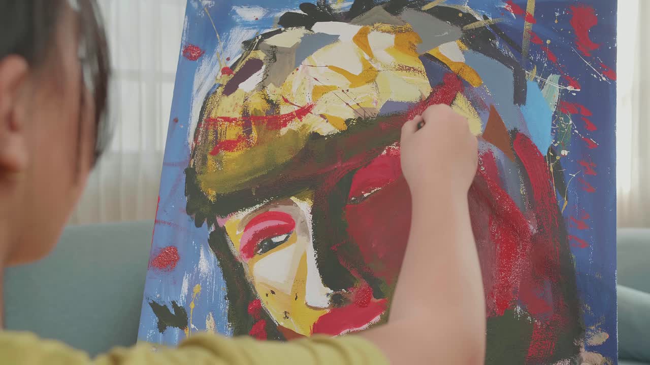 Close-up van een Aziatisch kunstenaresje dat op een ronde houten stoel zonder rugleuning zit, een penseel met gemengde kleuren vasthoudt en het gezicht van een meisje op het doek schildert