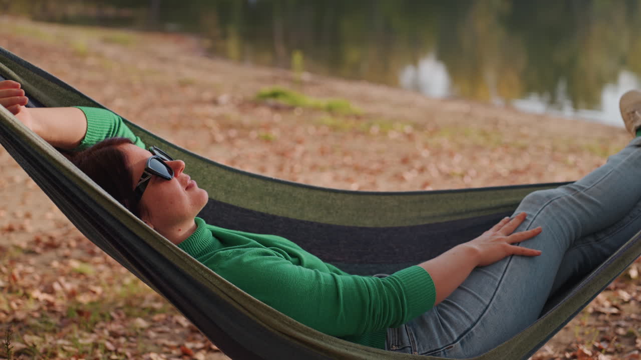 White woman in hammock by lake green sweater sunglasses jeans autumn leaves on ground calm lakeside campsite mood quiet reflection resting hands slow breath freelance writer taking nature break
