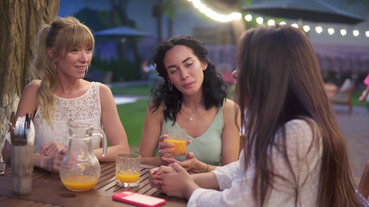 tres elegantes amigas se sientan en el café al aire libre, beben jugo y se divierten comunicándose