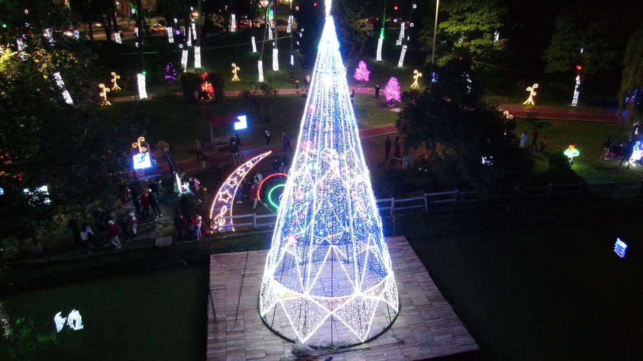 Big tree full with light and a bright shining peak in the illuminated by christmas lights bridge and groom park in Bogota. Drone dolly shot