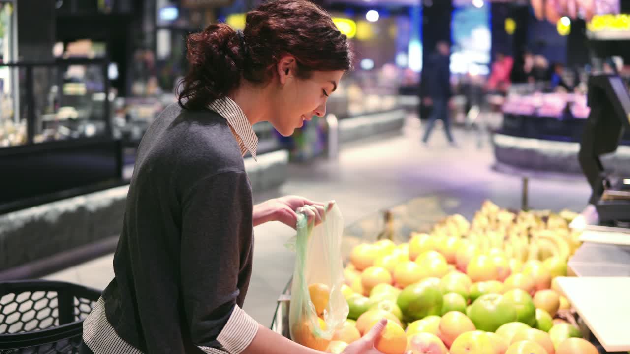 Young beautiful brunette girl in her 20's picking out oranges into a plastic bag at the fruit and vegetable aisle in a grocery store.