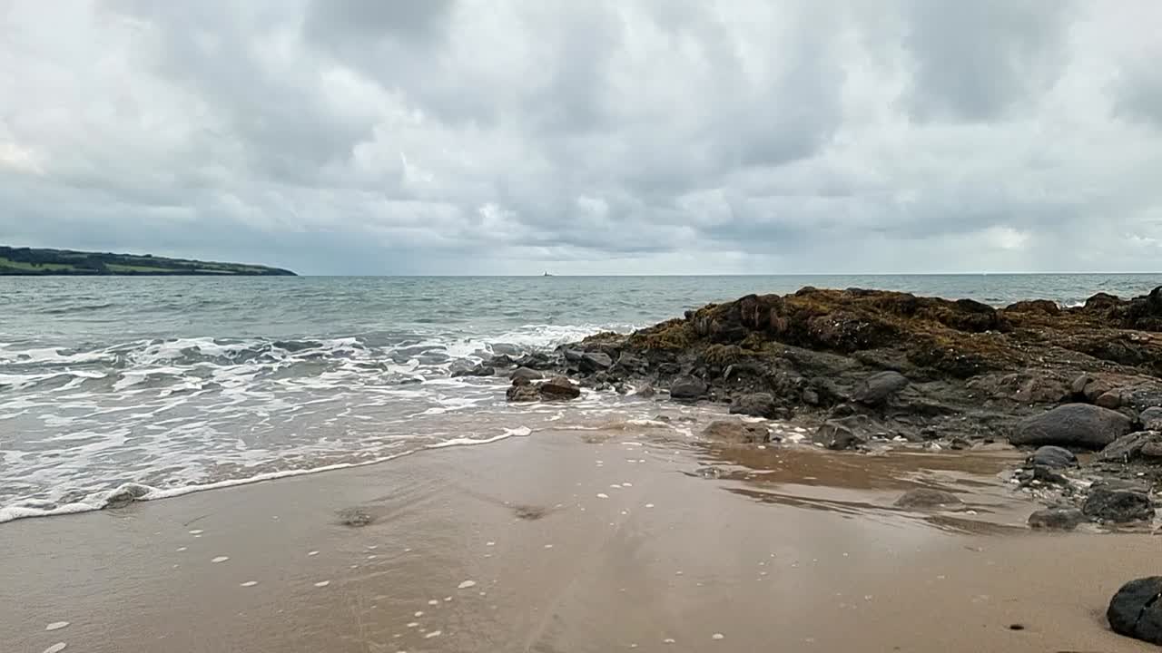 Calm slow motion ocean waves washing across Anglesey island splashing over rocks on golden sandy beach, overcast morning