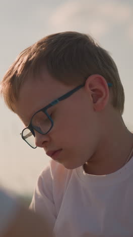 un primer plano de un niño con gafas y una camisa blanca, con una expresión reflexiva e infeliz en su rostro. su hermano está borroso en el fondo, añadiendo profundidad a la escena