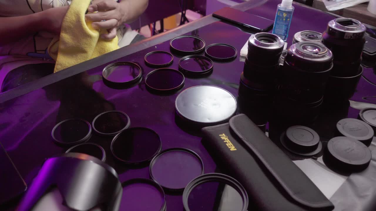 Person cleaning camera lenses and filters on a reflective table under purple lighting