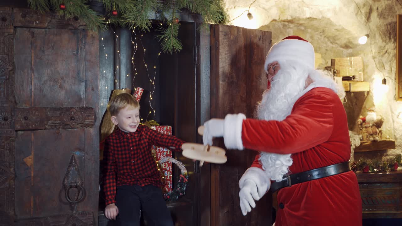 Santa Claus in red costume with white beard shows homemade toy plane to a little boy. Christmas present for obedient child. Cute boy looks at a wooden plane in Santa's hands.