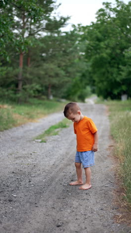 little male kid in orange t-shirt and shorts stepping carefully by the road with his bare feet. toddler is picking stones from the ground path. vertical video.