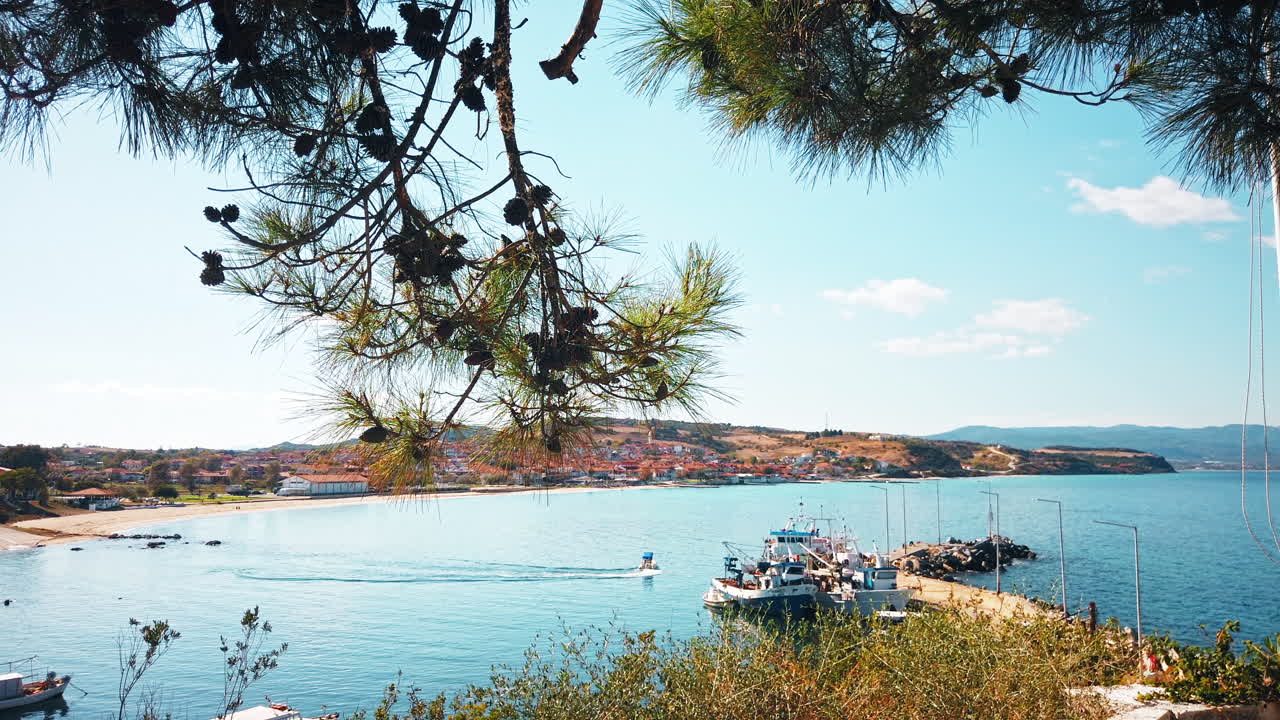View of a greek town through fir branches. Pier, moored and mooving boat, beach, buildings, hills