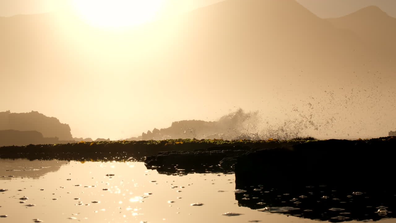 Splashing waves on rocky coastline backlit by bright golden sunrise, telephoto