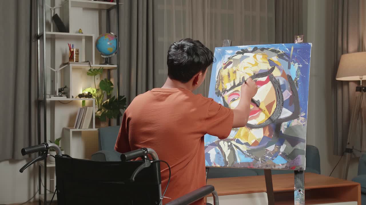 Hind View Of An Asian Artist Boy In Wheelchair Holding Paintbrush And Painting A Girl'S Face On The Canvas