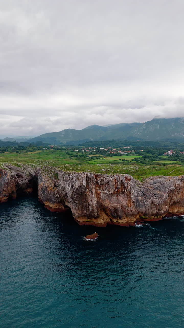 Spectacular view of the rocky cliffs on the shore of the sea. Picturesque village is located in the valley. Mountains covered with white clouds at backdrop. Vertical video.