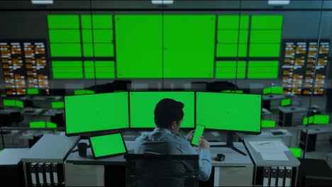 vista trasera de un hombre asiático usando un teléfono móvil y varios monitores de computadora con una pantalla verde simulada en la oficina
