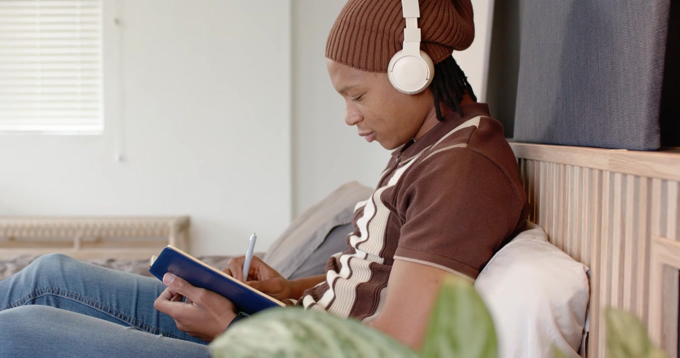 Man sitting on bed with blue notebook on lap starting writing with pen with headphones, journaling