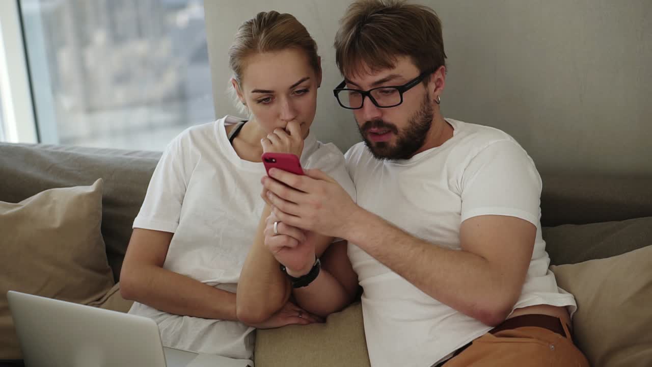 Attractive young couple watching smart phone together sitting on a couch in a living room at modern loft interior. Beautiful young caucasian couple using phone at home. Man showing something