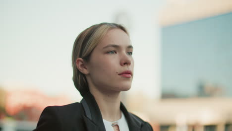 female stylist in black suit walking with confident expression and hair clipped back, looking ahead with blurred modern cityscape in background during bright workday morning in urban environment