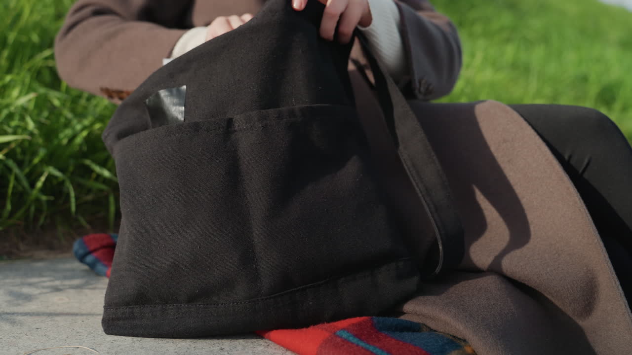 Persona su una panchina con una borsa nera, che indossa un cappotto marrone, aggiusta le cinghie e sistema gli effetti personali su una coperta colorata accanto a un bordo erboso, mani che chiudono la cerniera interna, movimento attento, atmosfera da parco urbano.