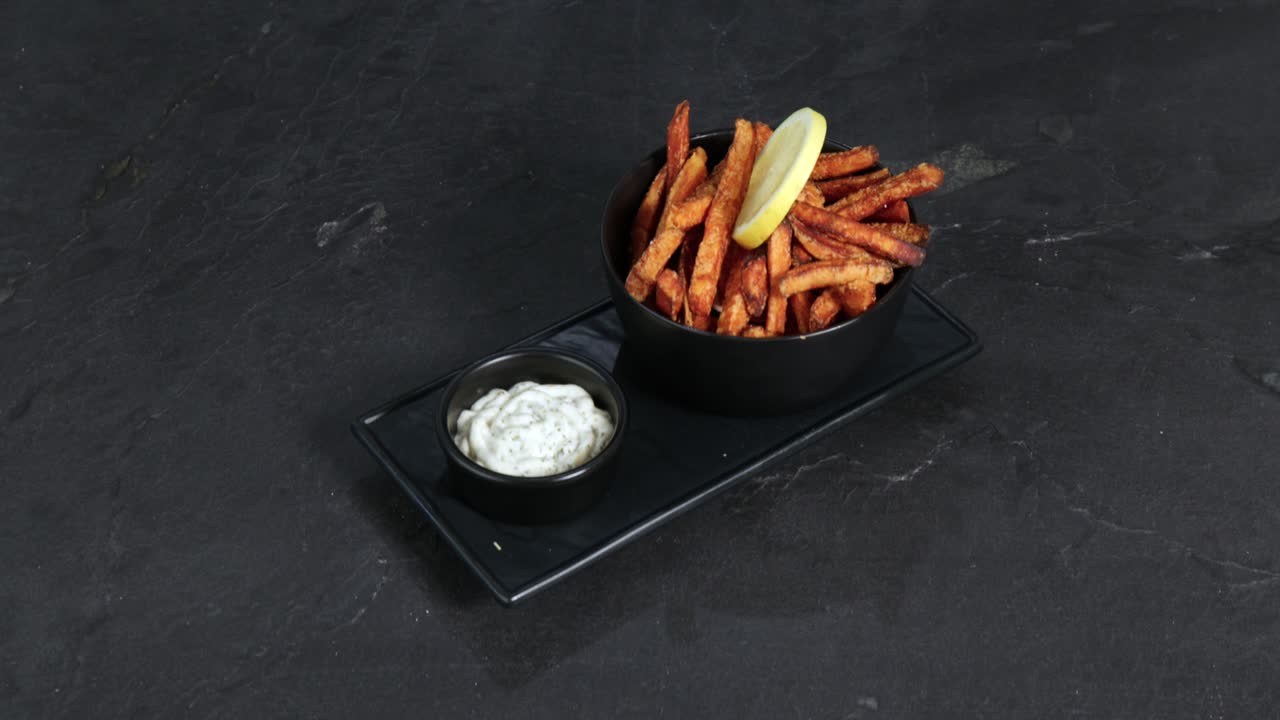 Sweet potato fries garnished with a slice of lemon next to a mug of mayonnaise are presented on a rotating plate