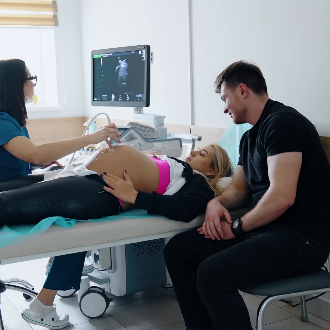 Una donna incinta è distesa sul letto mentre il medico le passa l'ecografo sulla pancia. Il marito, emozionato, è seduto accanto a lei e guarda il monitor.
