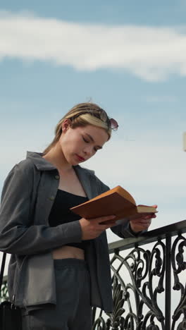 bela mulher de roupas cinzentas repousa sua mão direita em uma cerca de ferro enquanto lê um livro, o vento sopra suavemente seu cabelo e roupas, criando um momento sereno enquanto ela se concentra no livro