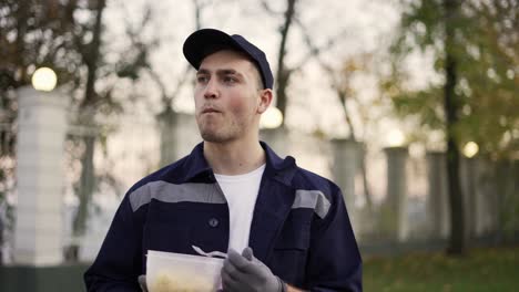 relaxed courier in blue uniform , enjoying taking food from lunchbox and standing in autumn street near the cardboard boxes