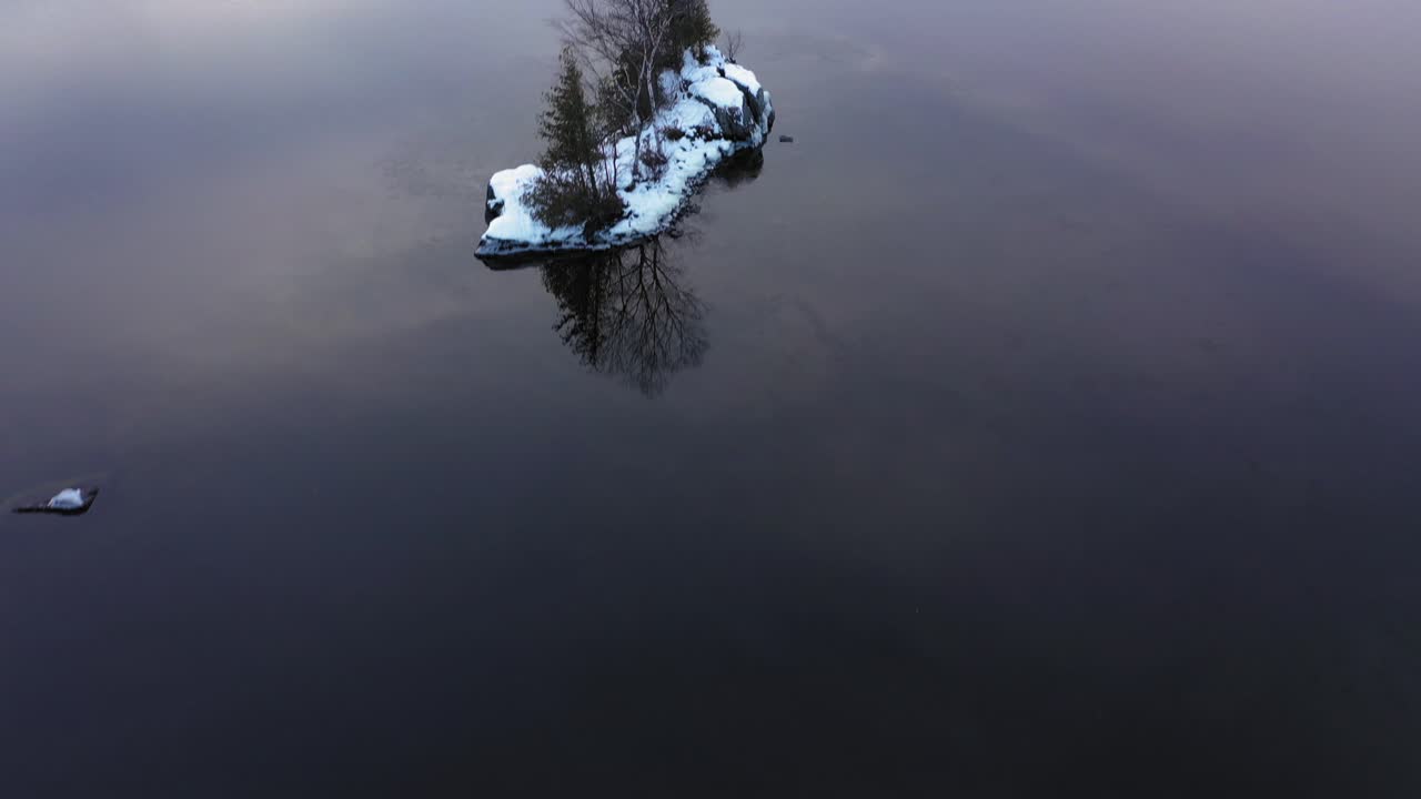 съемка с воздуха, пролетая низко над прозрачными спокойными водами, показывает скалистое дно и отражения облаков, когда проплывает небольшой покрытый снегом остров