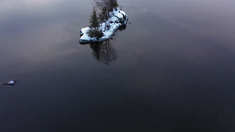 aerial footage flying low over the clear calm waters revealing the rocky bottom and cloud reflections as a small snow covered island is passed