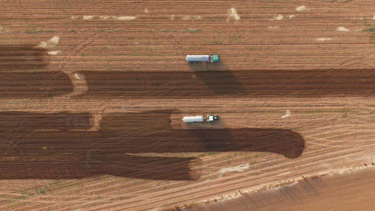 Multiple tractors spray manure in unison on large farmland in wide drone overhead top down tracking