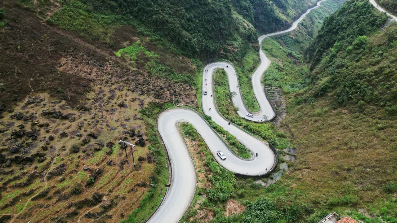 Pemandangan udara jalan pegunungan berkelok-kelok dengan tikungan tajam dan kelokan lebar melintasi perbukitan hijau terjal saat kendaraan bergerak melalui pemandangan dramatis di Ma Pi Leng Pass, Ha Giang