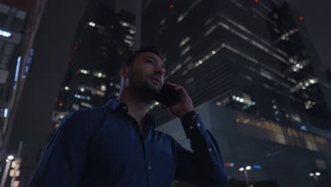 Businessman-picks-up-a-smartphone-call-and-talk-on-phone-near-office-at-night,-successful-employee-standing-near-skyscraper-building-talking-on-cellphone