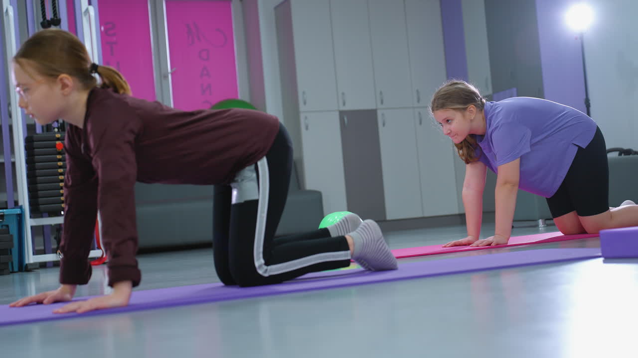 vista lateral de chicas jóvenes realizando ejercicios de flexibilidad de la columna vertebral en esteras en un gimnasio, con la chica de púrpura observando atentamente mientras la luz suave ilumina el fondo