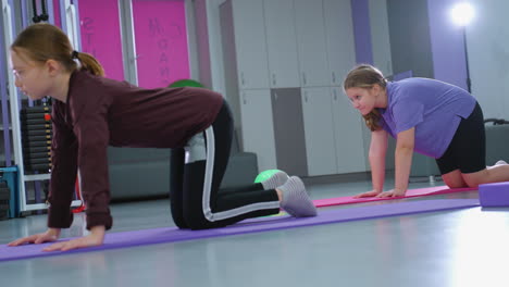 side view of young girls performing spine flexibility exercises on mats in a gym, with the girl in purple watching intently as soft light illuminates the background