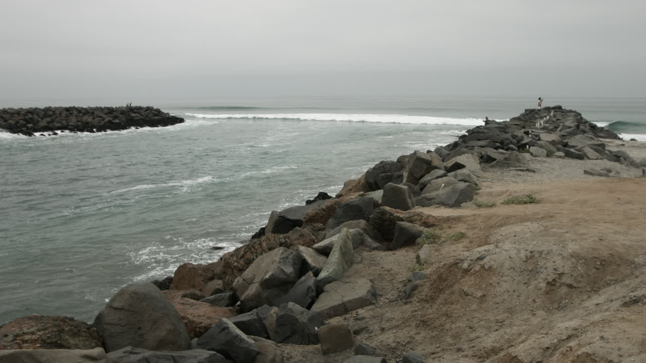 Cinematic UHD footage of the ocean at a quiet inlet in Carlsbad, California, on an overcast morning. Gentle waves roll in under grey skies, creating a calm, moody, and reflective coastal scene.