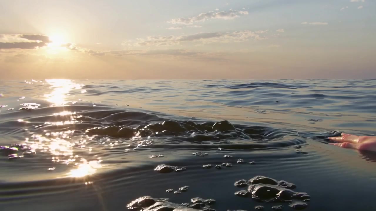 Mão nua aparecendo da borda direita, roçando a superfície da água ao pôr do sol, criando ondulações e espuma.