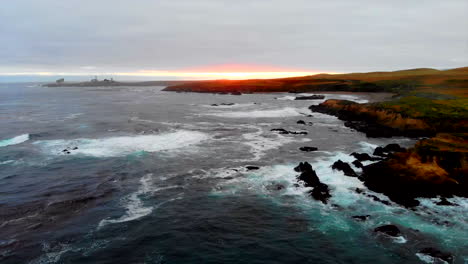 Pacific-Ocean-drone-booms-down-from-sky-to-water-level-of-gentle-waves-at-sunset-with-lighthouse-in-distance-on-Central-California-Coast-near-Hearst-Castle,-Highway-1,-and-Big-Sur,-4k-Pro-Res-422-HQ