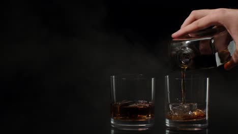 barman pouring whiskey, cognac, brandy from bottle into glasses with ice cubes on black background