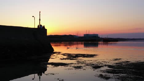 Swans-glide-across-placid-water,-stunning-pink-and-orange-sunrise-reflect,-locals-take-in-views-at-Nimmos-Pier-Galway-Ireland