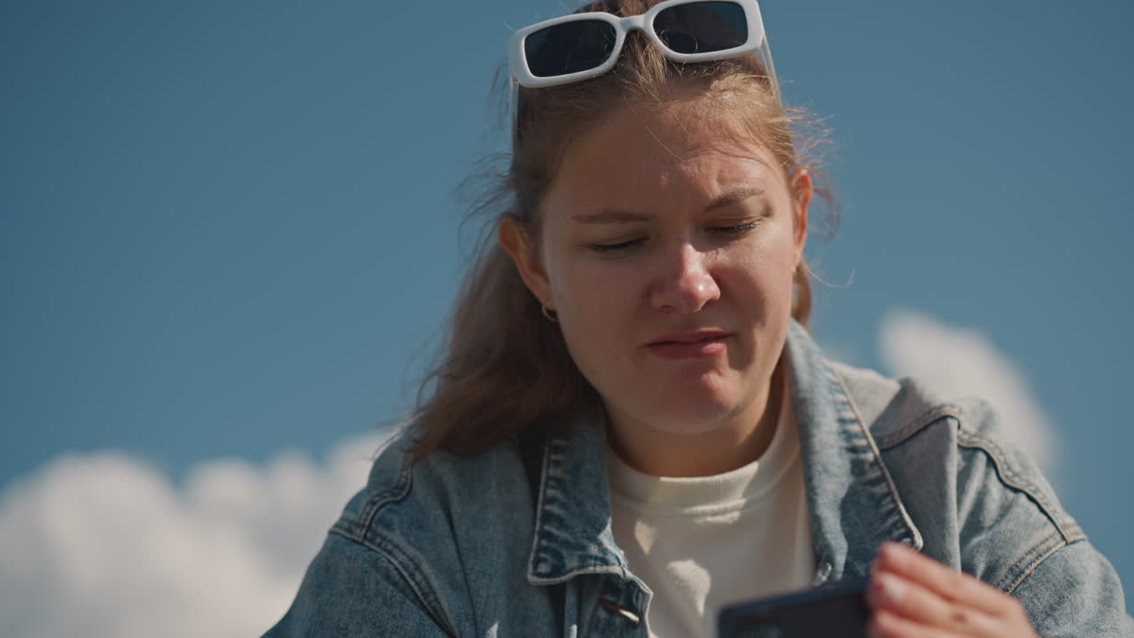 Kaukasisk kvinne undersøker telefonen med bekymring, nærbilde av ansiktsuttrykk, dongerijakke, solbriller på hodet, overskyet blå himmel i bakgrunnen, subtil frustrasjon, forsøker reparasjon eller innstillinger, naturlig.