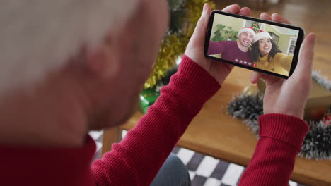 hombre caucásico con sombrero de santa usando un teléfono inteligente para una videollamada de navidad, con una pareja en la pantalla