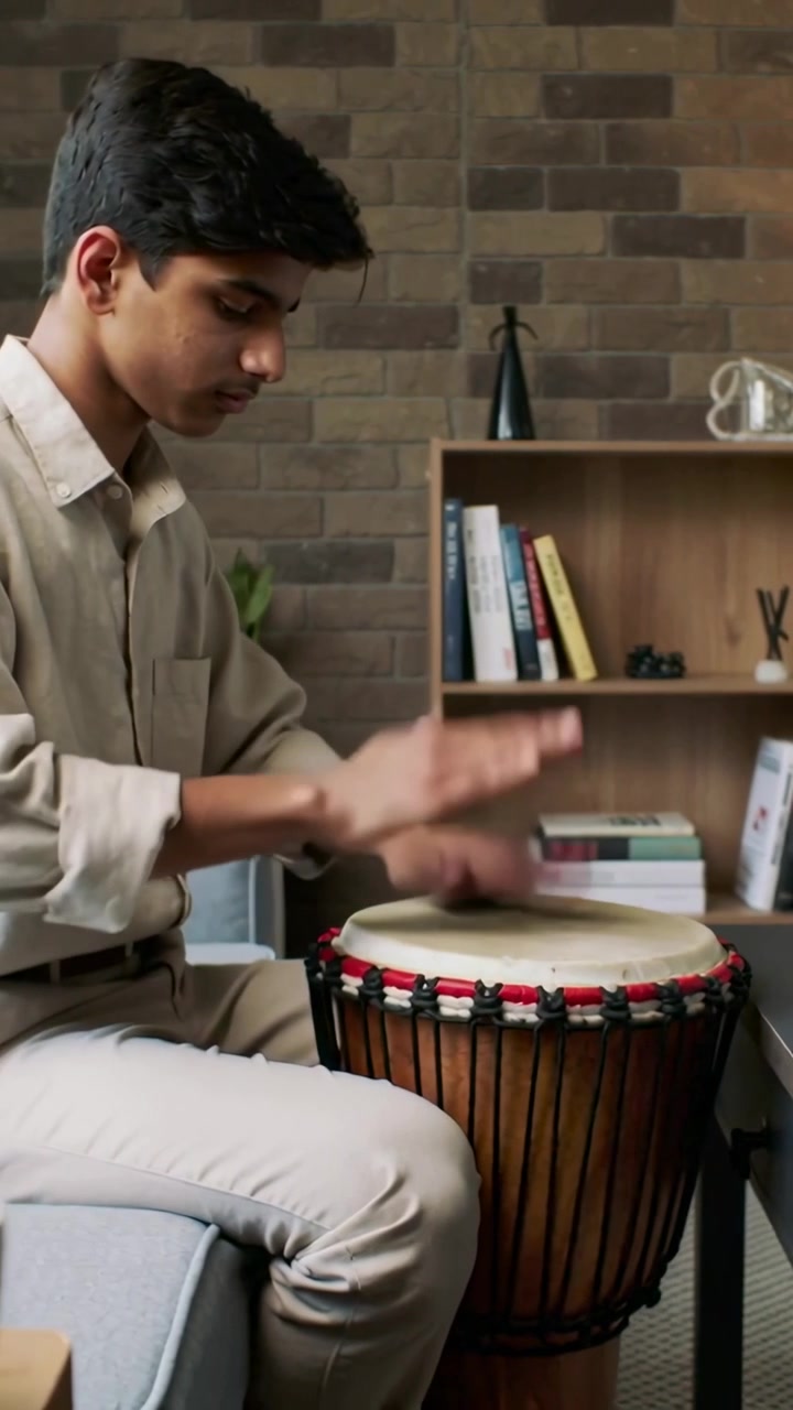 Video vertical: Cámara grabando, adolescente indio vestido de beige tocando un djembe en casa, practicando junto a un portátil.