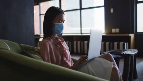 asian woman wearing face mask using laptop while sitting on sofa couch at modern office