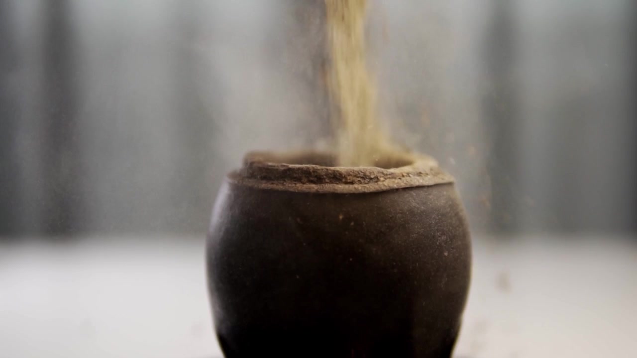 Close view of loose tea or dried herbs pouring into traditional dark ceramic vessel. Fine particles fall steadily against the soft blurred background