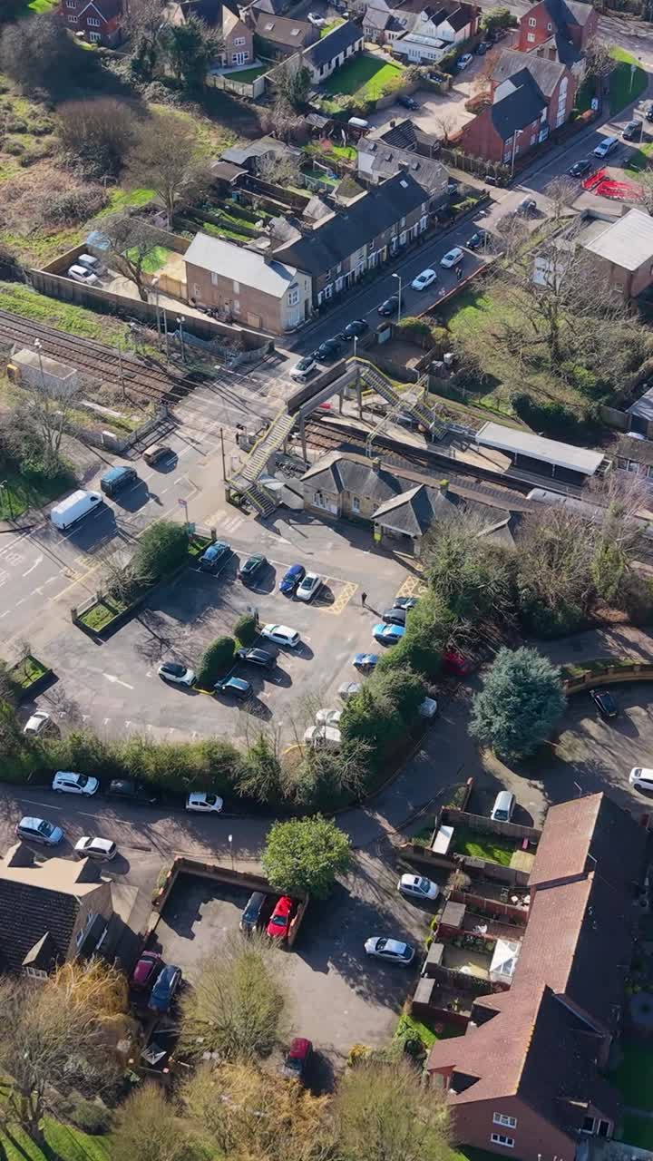 Un treno arriva alla stazione di Stanstead St Margarets mentre un drone orbita a sinistra sopra un passaggio a livello chiuso con auto in attesa, mostrando i binari, i marciapiedi e le strade del villaggio nell'Hertfordshire. Verticale.
