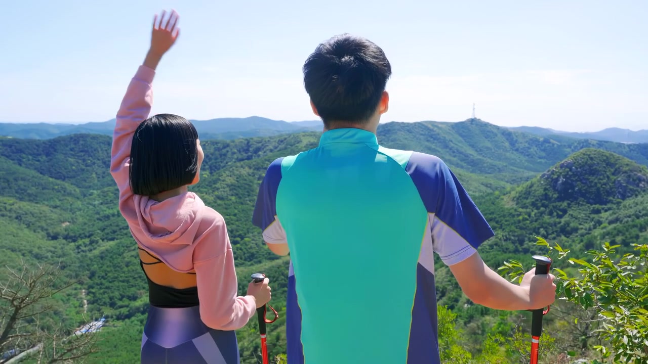 カップルのバックパッカーがトレッキングウェアを着用し、山頂に立っている。山頂到達を祝い、勝利を喜びながら両腕を広げ、手を高く掲げて興奮気味に飛び跳ねている。