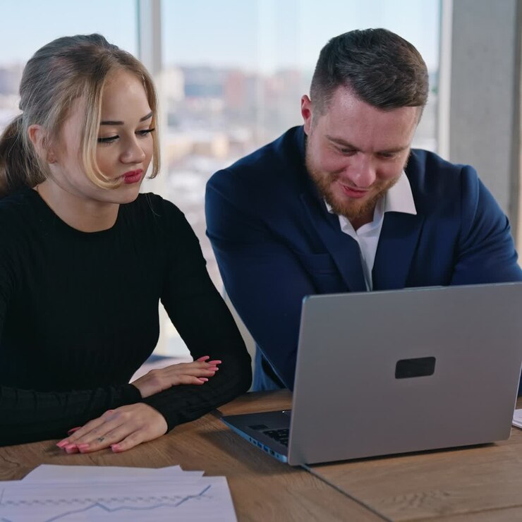 Young pretty collegues work on business. Beautiful female and young businessman sitting together at the table and look into a laptop in office room on city window background