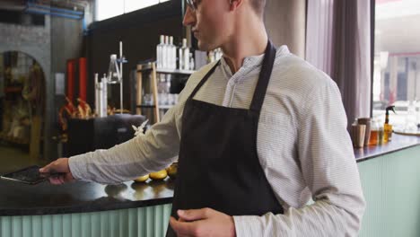 portrait of caucasian man wearing apron working at a bar, using tablet and smiling to camera