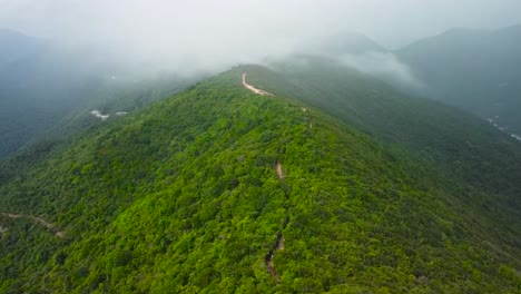 aerial drone footage rising up over a mountain in china asia called unicorn ridge while white fluffy clouds and mist is hovering in between the mountains. a dirt muddy brown road going up the hillside