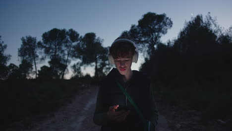 teenage male walking isolated dirt path at twilight, listening to music on smartphone amid shadowy rural landscape and dimming forest treeline