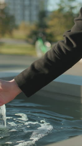 close up of man in black coat multitasking with tablet near spring water outdoors, hand poised for interaction, bright sunlight illuminating scene with blurred urban greenery