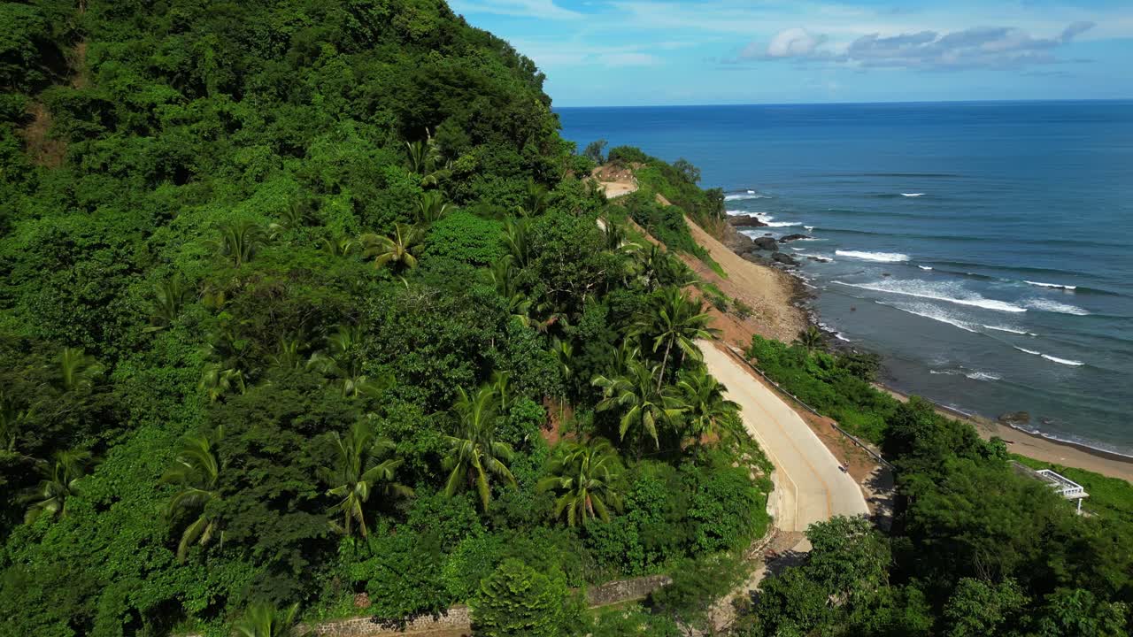 Swivel pan aerial of Dingalan, Aurora, showcasing lush green trees and the winding hillside road carved into the slopes, with turquoise sea and waves framing the dramatic coastline