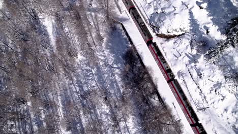 Aerial-view-of-the-Bernina-Express-panorama-train-going-through-a-snow-covered-mountain-winter-landscape-with-forests-on-a-sunny-day-in-Alp-Grum,-Switzerland