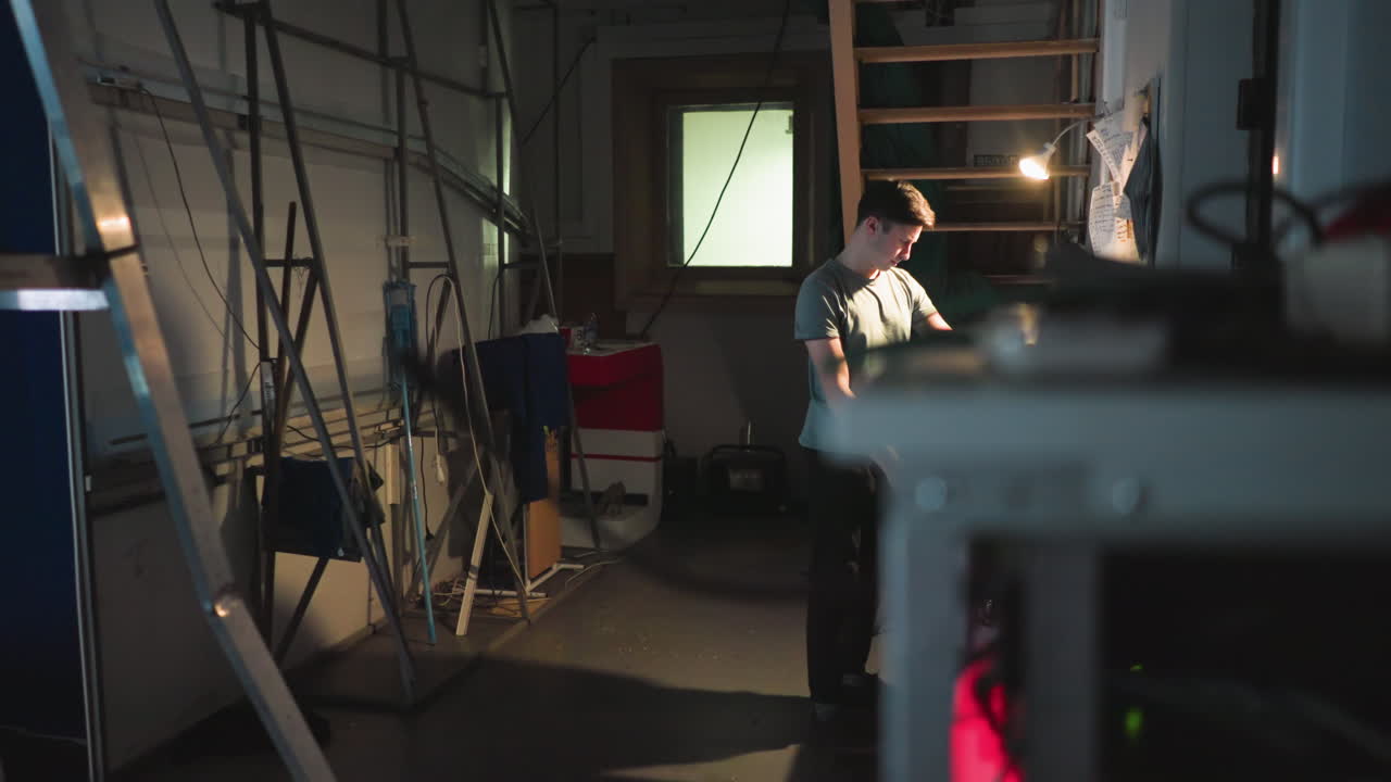 Man operates illuminated touchscreen control panel in dim industrial studio workspace filled with ladders, cables, and technical equipment, standing near wall with notes